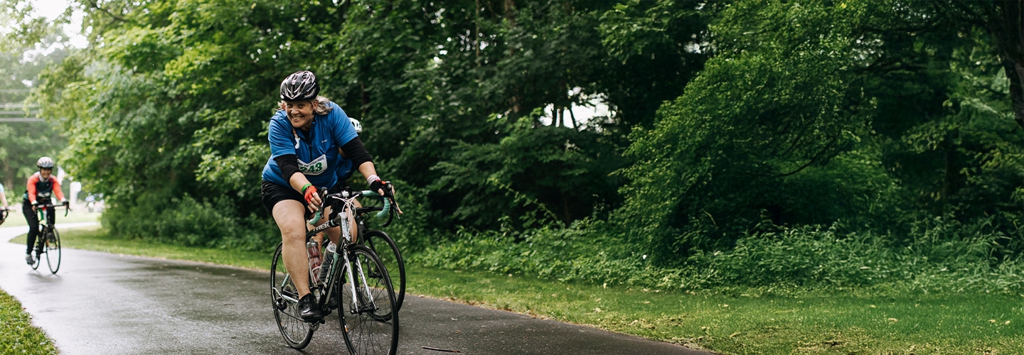 Smiling cyclist wearing a helmet and blue shirt with a race bib rides a road bike on a greenway trail, with other cyclists following behind.